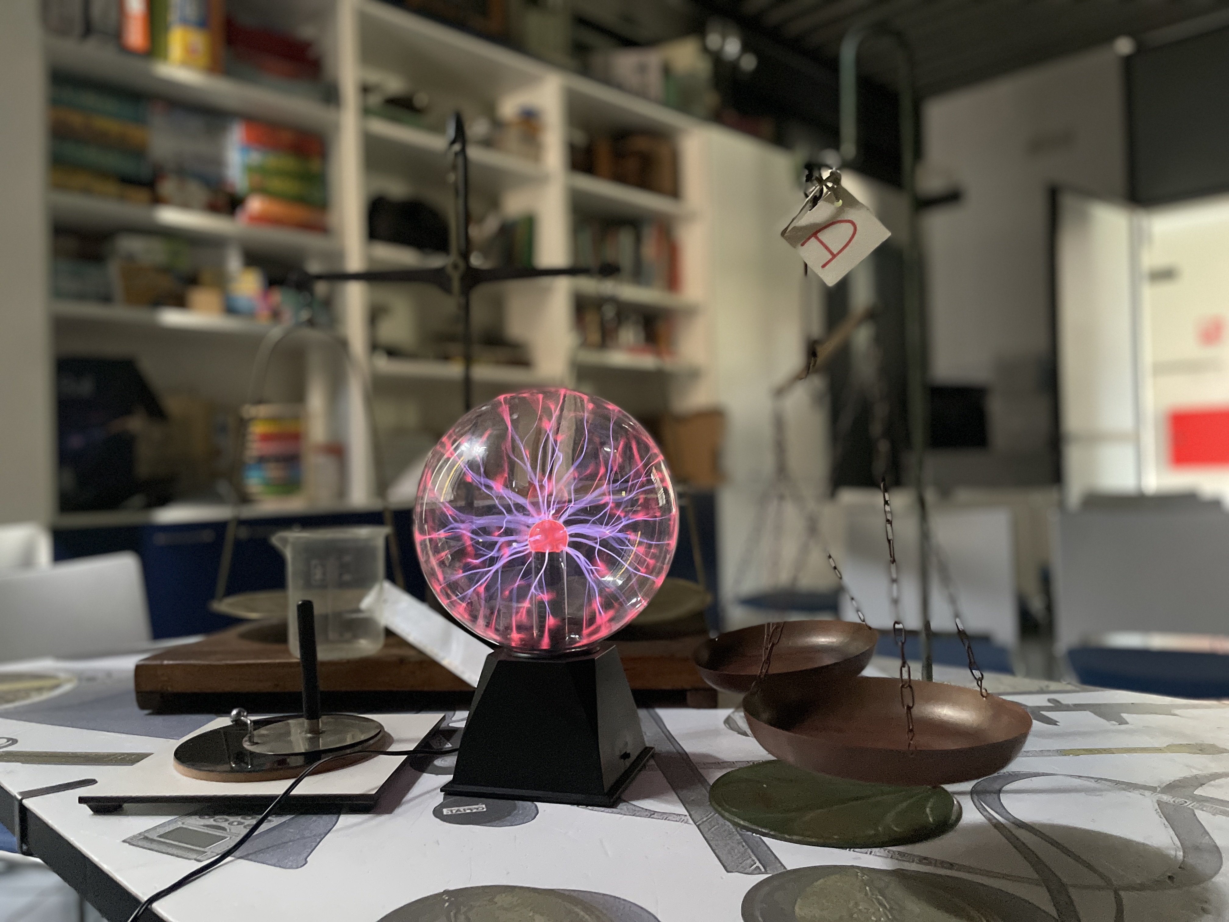 Sul tavolo, di una sala espositiva del museo, c'è una lampada a sfera al plasma con i raggi di energia fucsia e viola. Accanto una vecchia bilancia con i due piatti e alcuni strumenti di misurazione.