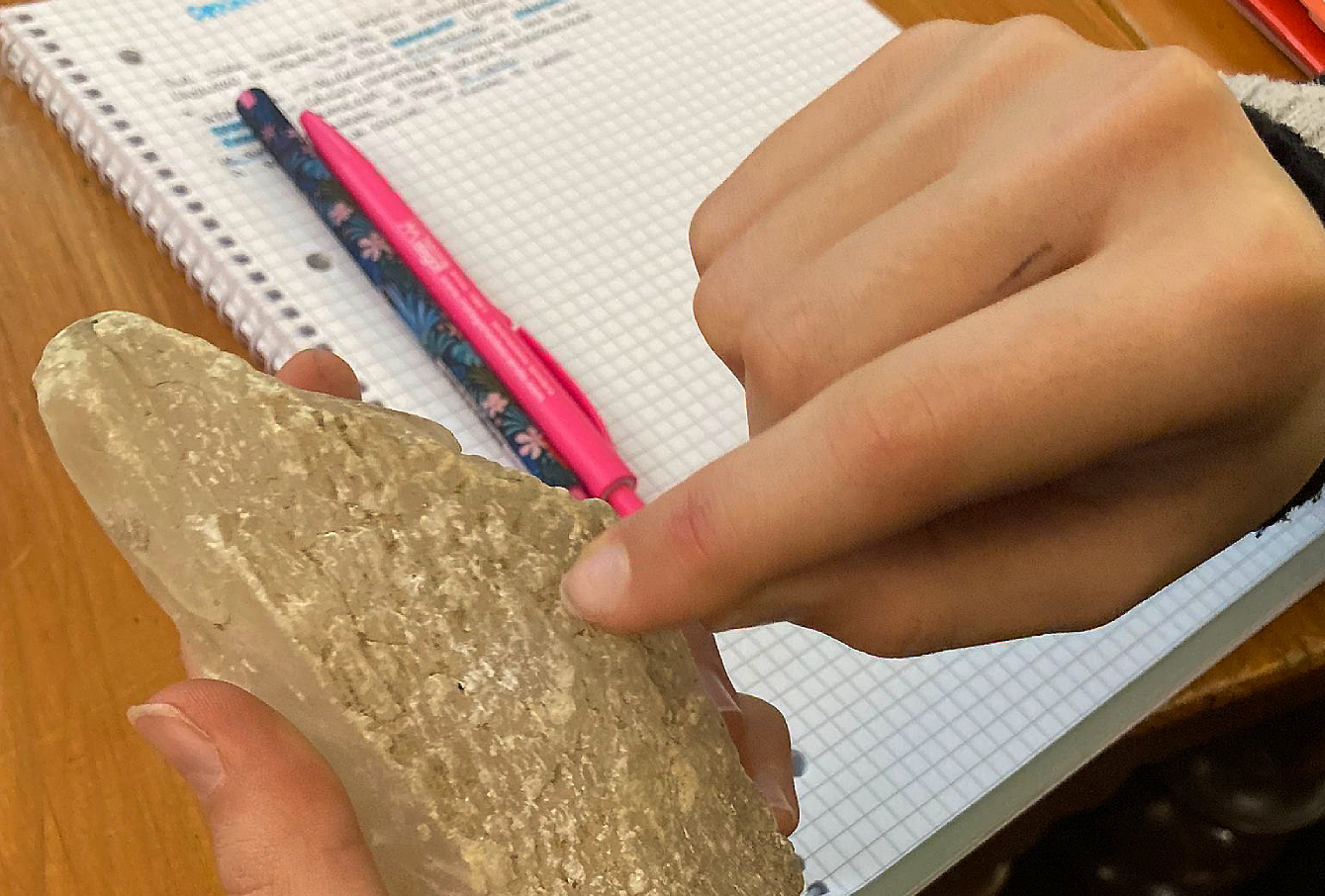 Osservare le rocce - Le mani di un bambino reggono e toccano una roccia . Sullo sfondo un quaderno scolastico su cui annotare le informazioni sensoriali raccolte, con una penna e una matita.
