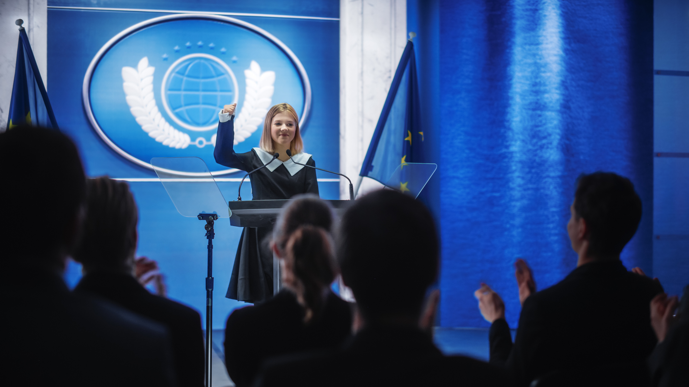 Foto di una bambina con accanto la bandiera dell'Unione Europea mentre sul palco parla al pubblico ripreso di spalle. 