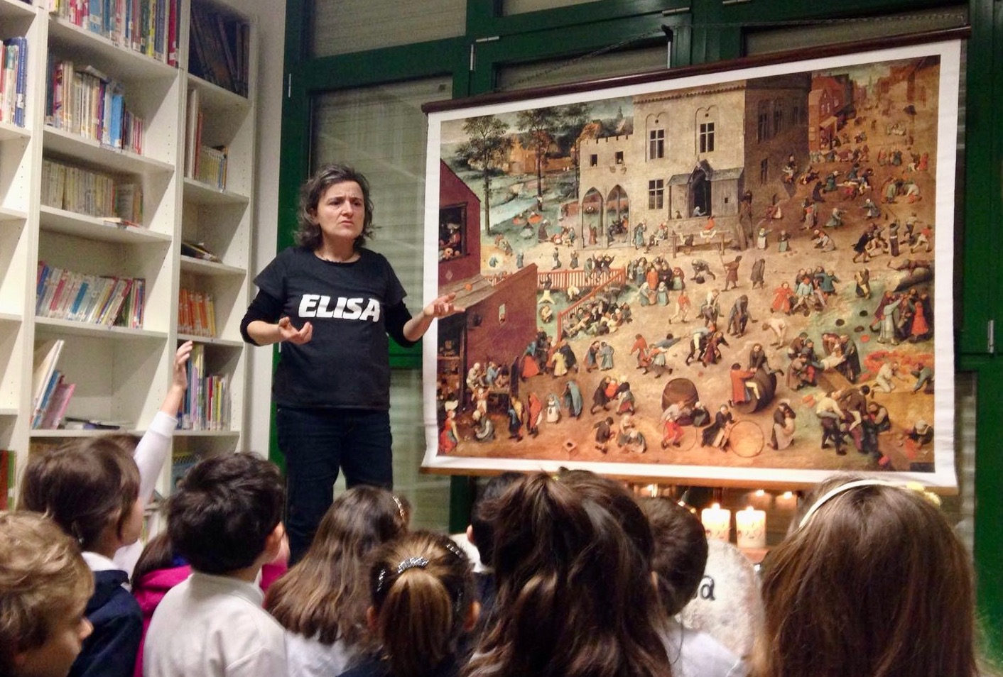 Elisa in una stanza mostra il quadro "Giochi di bambini" di Pieter Bruegel, a grandezza reale di 130x160 cm, ad una classe di bambini seduti per terra